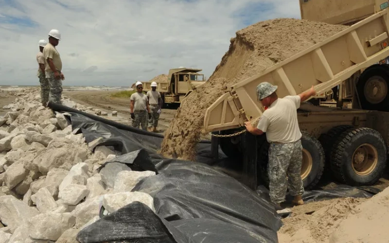 army workers watching a dumptruck pouring dirt on top of an area with geotextile